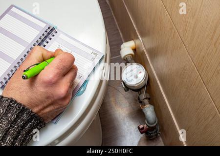 Male hand writing water consumption from a water meter counter inside a apartment bathroom,  close up Stock Photo