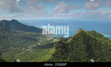 Aerial view of tropical island with mountain ridge covered in green rainforest , turquoise lagoon, coral reef, breathtaking landscape under cloudy blue sky. Remote wild paradise, exotic summer travel Stock Photo