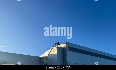 A mostly clear blue sky above a large, light gray building under construction. A cherry picker is positioned on the building's upper section.  The bui Stock Photo