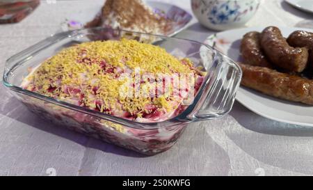 A glass dish of layered beet salad topped with shredded egg yolk sits on a table next to a plate of cooked sausages.  Other food items are visible in Stock Photo