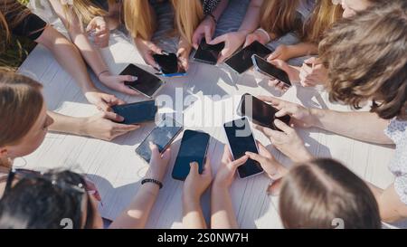 High school students enjoying summer outdoors at a white table, using smartphones to connect through social media Stock Photo