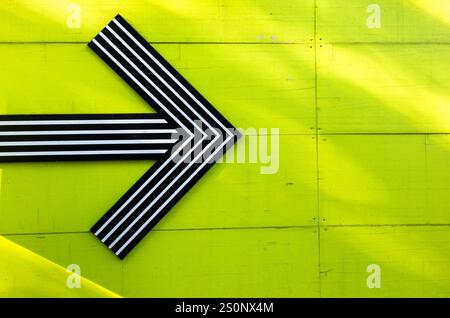 A bold black and white striped arrow affixed to a vibrant lime green wooden wall. The arrow points to the right, suggesting direction or movement. Stock Photo
