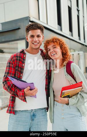 Vertical. Two cheerful classmates posing together outdoors, smiling warmly at the camera while holding books and backpacks, showcasing a friendly and happy student life outside their university campus. High quality photo Stock Photo