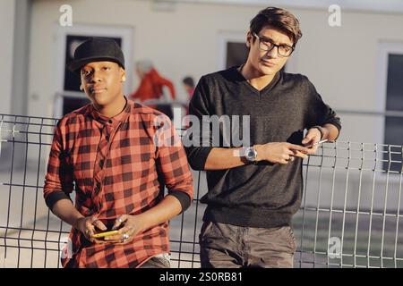 Two young adult men leaning casually against a metal fence, engrossed in their smartphones while enjoying a relaxed moment outdoors in an urban settin Stock Photo