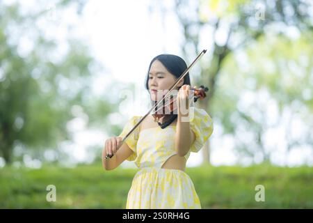 Twenty-something Chinese woman in yellow dress gracefully playing violin in the forest of a large, nature-filled park in Bukit Jalil, near Kuala Lumpu Stock Photo