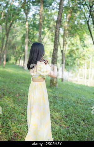 Twenty-something Chinese woman in yellow dress gracefully playing violin in the forest of a large, nature-filled park in Bukit Jalil, near Kuala Lumpu Stock Photo