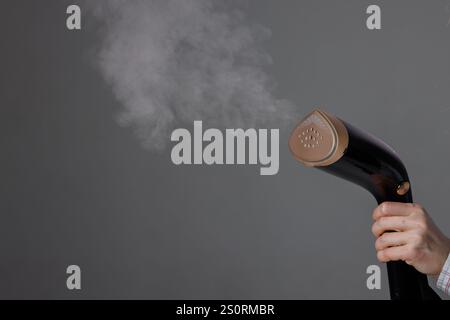 handheld steamer in woman's hand with steam on grey background, close up. Stock Photo
