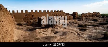 A panoramic view of ancient ruins with a mud-brick wall in the background. The landscape features rocky formations and sparse vegetation under a clear Stock Photo