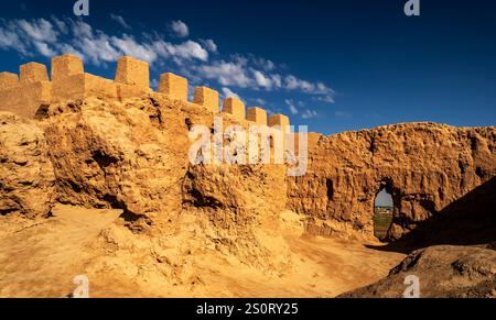 A historic fortress made of mud bricks, featuring a large archway and a wall with battlements. The landscape is arid with a clear blue sky and scatter Stock Photo