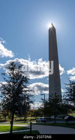 The Washington Monument stands tall against a bright blue sky with fluffy clouds, sunlight shining from behind its peak, surrounded by green trees and Stock Photo