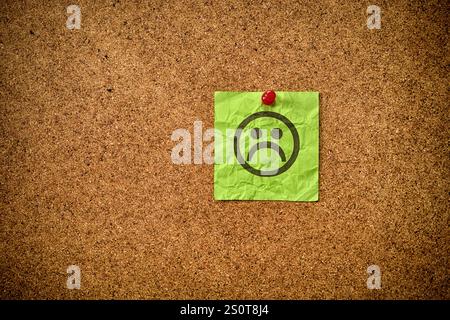 A green sticky note with a sad face on it pinned to a corkboard (bulletin board). Close up. Stock Photo