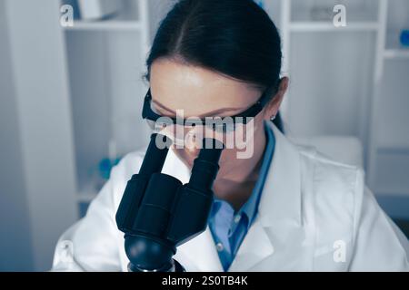 In a modern lab, a dedicated female scientist analyzes samples under a microscope, demonstrating her expertise and commitment to research and Stock Photo