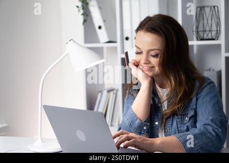 A focused woman is diligently working on her laptop in a bright and inviting home office environment, showcasing a perfect blend of creativity and Stock Photo