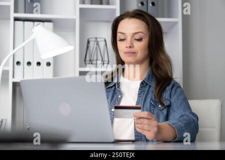 A determined and focused woman is shopping online using her credit card at her desk, comfortably engaged in the vast digital marketplace, which Stock Photo