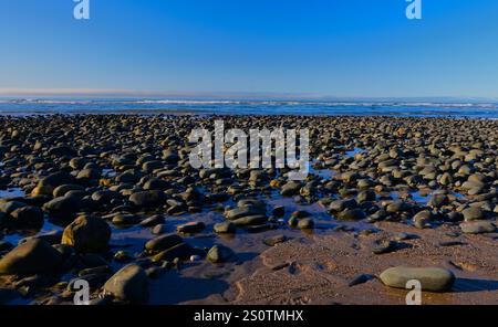 Pebbles on beach, big and small with small waves in background under a blue winter sky. Stock Photo