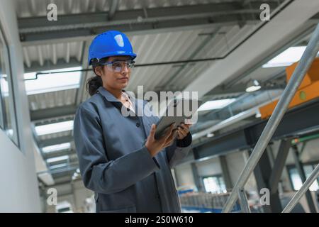 Young Indian woman with a blue helmet, an engineering programmer at a workplace in a factory, looking at graph data on a tablet device. Stock Photo