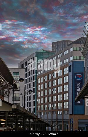 Modern city buildings under a vibrant, colorful sky.  A Travelodge sign is visible on one building.  The image features architectural details and a tr Stock Photo