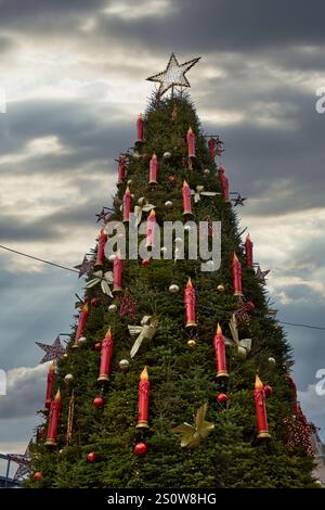 Large Christmas tree decorated with numerous red candles, gold bows, and stars under a cloudy sky.  The tree is tall and densely foliated in Birmingha Stock Photo