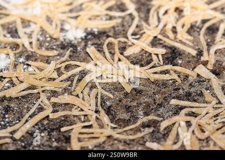 Close-up view of uncooked, pale yellow pasta strands scattered on a dark brown, textured surface with visible flour dust.  The pasta is thin and irreg Stock Photo