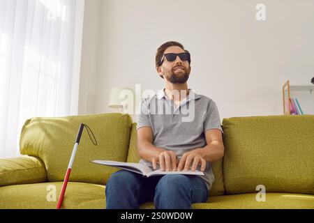 Blind man in dark eyeglasses sitting on sofa at home and reading tactile braille book. Stock Photo
