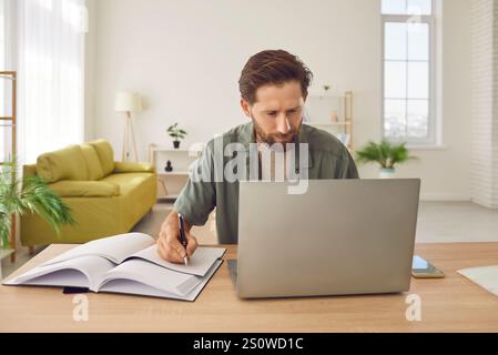 Man working at the desk with a laptop at office or at home making notes in his notepad Stock Photo