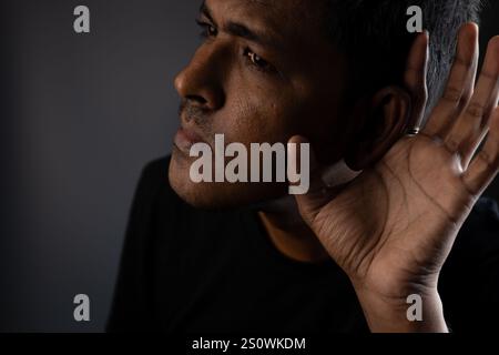Man cups hand to ear, straining to hear a faint sound in the shadows.  A portrait of focused listening and anticipation. Stock Photo