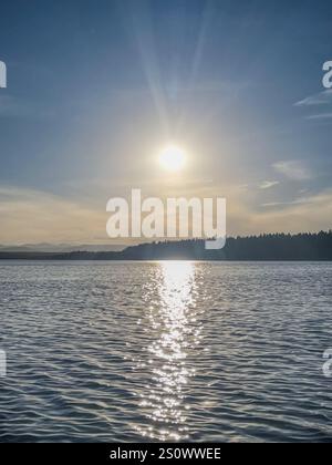 Scenic view of a serene lake with sunlight reflecting on the water during dusk, framed by distant forested hills and a clear blue sky. Stock Photo