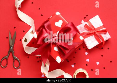 Beautiful gift boxes with ribbon, scissors and confetti on red background. Valentines Day celebration Stock Photo