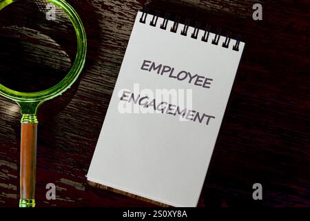 A notepad with the bold text Employee Engagement sits on a wooden surface beside a magnifying glass, symbolizing the focus on workplace morale and pro Stock Photo