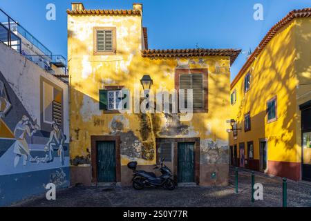 A charming urban scene featuring a vibrant yellow-painted building with weathered details, a nearby mural depicting figures, and warm sunlight casting Stock Photo