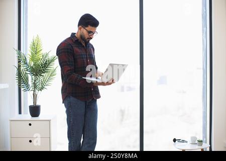Indian man standing near large window using laptop in modern office. Mid-adult male in plaid shirt focused on work. Bright and spacious environment with natural light and minimal decor. Stock Photo