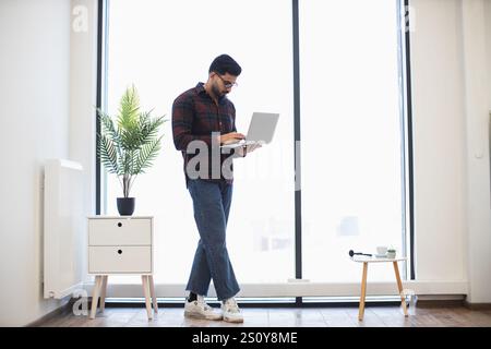 Young Indian male standing in bright office using laptop, showcasing modern work environment. Features include plant decor, wooden furniture, large window. Stock Photo