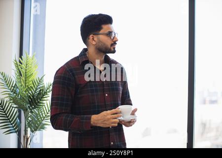 Indian man in his 30s wearing glasses holds coffee cup by window. He appears thoughtful and relaxed in casual environment with indoor plant adding natural touch. Stock Photo
