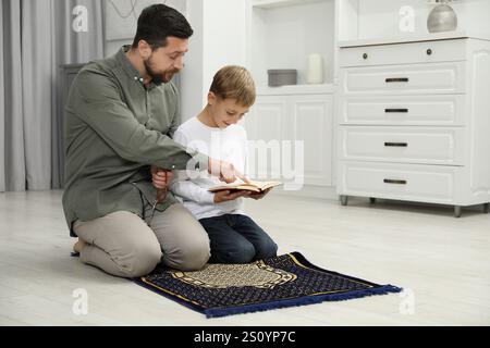 Muslim man with his son reading Quran and praying on mat at home, space for text Stock Photo