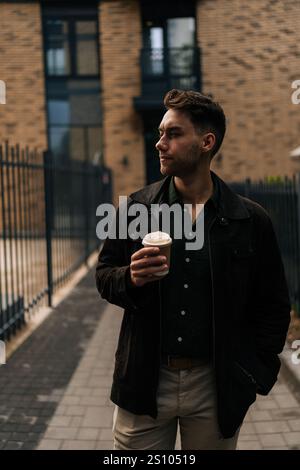 Vertical portrait of confident young man pensive looking away while holding coffee cup, enjoying drink standing in urban alleyway by brick buildings Stock Photo