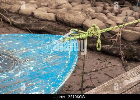 The bow of a small blue boat is placed on a muddy shore, where rocks are also visible. The surface of the boat is worn and the paint has peeled off in Stock Photo