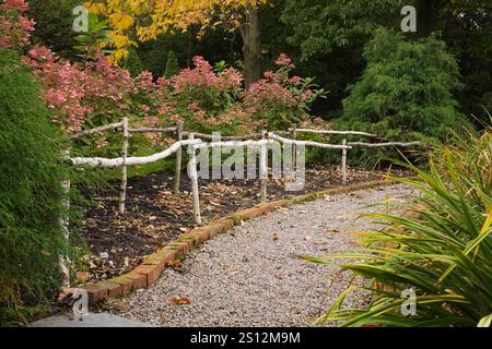 Gravel path through borders with Hydrangea paniculata ‘Quick Fire’ shrubs, Hemerocallis stalks - Daylily and edged with red bricks and rustic railing. Stock Photo