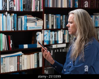A side-profile view of a man with long hair, deeply focused on his e-book, with a background of a bookshelf filled with books, creating a tranquil and Stock Photo