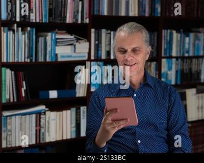 With a cheerful expression, a man wearing a blue shirt and a ponytail enjoys his e-book, surrounded by books on a cozy shelf. Stock Photo
