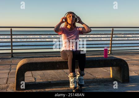 Young athlete woman wearing inline skates is sitting on a bench by the sea at sunset, listening to music with headphones and holding a pink bottle of Stock Photo