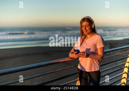 Young sportswoman with headphones is using her smartphone and listening music near the beach at sunset, enjoying the moment of relax after inline skat Stock Photo