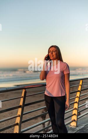 vertical Young sportswoman wearing pink t-shirt and black leggings talking on the phone while enjoying the sunset by the sea, leaning on the railing o Stock Photo