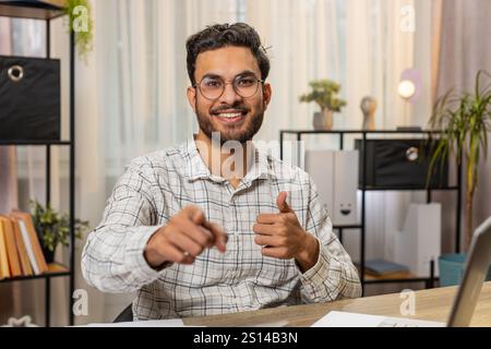 I choose you. Young Hispanic businessman programmer software developer working on office laptop pointing to camera looking with happy expression, making choice showing direction. Indian freelancer guy Stock Photo