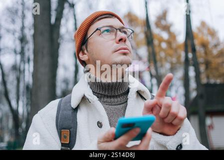 Young man wearing eyeglasses and warm clothing using smartphone and pointing finger while exploring park during autumn season Stock Photo