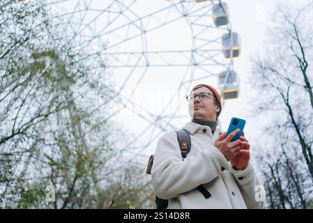 Young man with backpack and smartphone is standing near ferris wheel in amusement park, looking away and using mobile phone, wearing warm white jacket, orange hat and eyeglasses Stock Photo