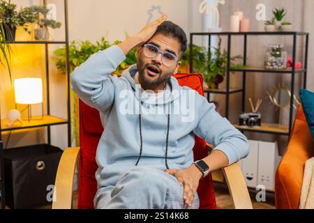 Face palm. Shame on you. Upset Indian man making disrespect gesture, feeling bored, disappointed in fail results, bad news. Unhappy confused Arabian guy at home apartment room sitting on rocking chair Stock Photo