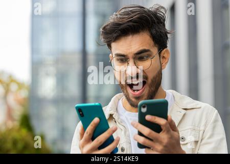 Irritated nervous stressed Indian man talking screaming on two mobile phones having conversation conflict quarrel complaint dispute discuss solve problem outdoors. Arabian upset guy in downtown city Stock Photo