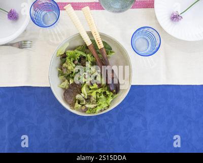 Top view of green salad served on the table with blue tablecloth and glassware. White plates decorated with fresh purple flowers Stock Photo