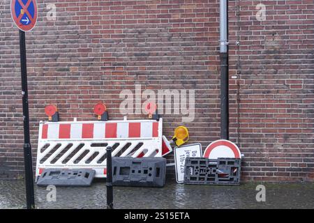 Red and white construction barrier, traffic signs, and bases stacked against a brick wall with no parking sign Stock Photo