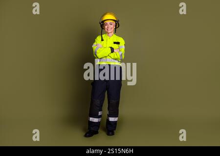 Portrait of a mature female worker in a yellow safety helmet and protective workwear, standing confidently against a khaki background Stock Photo